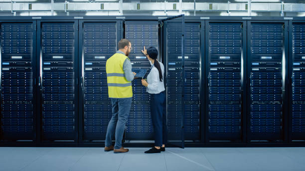 istock Bearded IT Admin in Glasses and High Visibility Vest with a Laptop Computer and Young Technician Colleague Talking in Data Center while Standing Next to Server Racks. Running Diagnostics or Maintenance. 1131198063