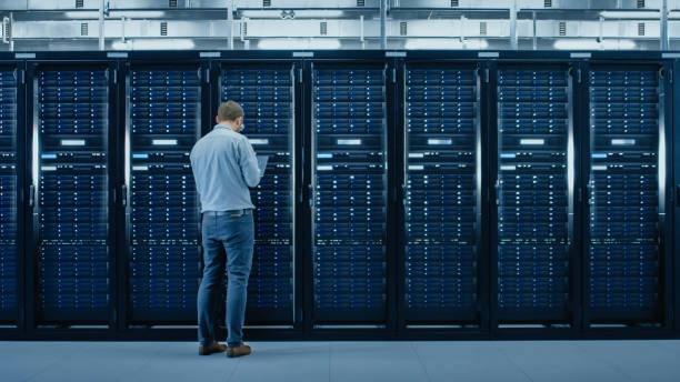 istock Bearded IT Specialist in Glasses is Working on Laptop in Data Center while Standing Before Server Rack. Running Diagnostics or Doing Maintenance Work. 1131198071
