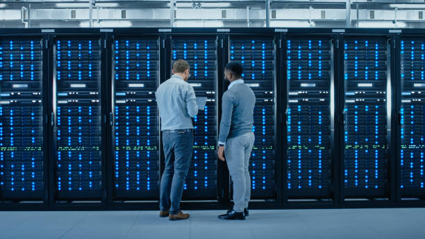 istock Bearded IT Engineer in Glasses with a Laptop Computer and Black Technician Colleague Talking in Data Center while Standing Next to Server Racks. Running Diagnostics or Maintenance. 1131198072