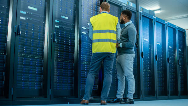 istock IT Administrator with a Laptop Computer in High Visibility Vest and Black Male Engineer are Talking in Data Center while Standing Before a Server Rack. Running Diagnostics or Doing Maintenance Work. 1131198078
