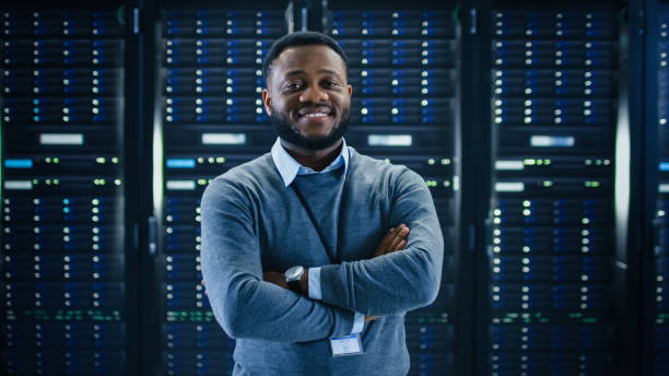 bearded black it engineer standing and posing with crossed arms in the middle of a working data center server room with server computers working on a rack. - network server rack computer black imagens e fotografias de stock