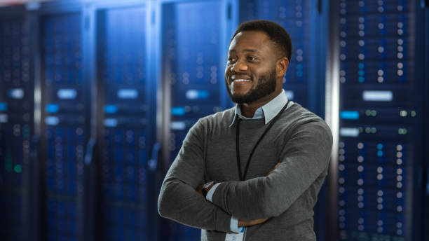 black data center it technician standing in the middle of a server rack corridor. he is standing with crossed arms and is smiling. - network server rack computer black imagens e fotografias de stock