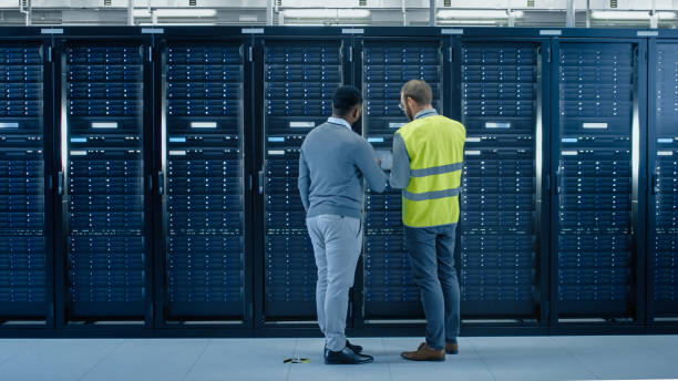 istock Bearded IT Specialist in Glasses and High Visibility Vest with a Laptop Computer and Black Technician Colleague Talking in Data Center while Standing Next to Server Racks. Running Diagnostics or Maintenance. 1131198101