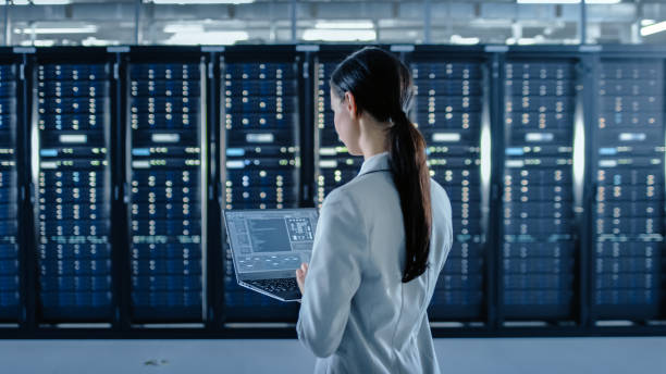istock Back Shot of a Female Data Center IT Specialist Works on Laptop Computer Next to Server Rack Corridor. Running Diagnostics or Doing Maintenance Work. 1131198109