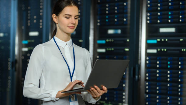 istock Beautiful Data Center Female IT Technician Standing Near Server Rack Corridor with a Laptop Computer. She is Using Notebook and Working with Servers. 1131198125