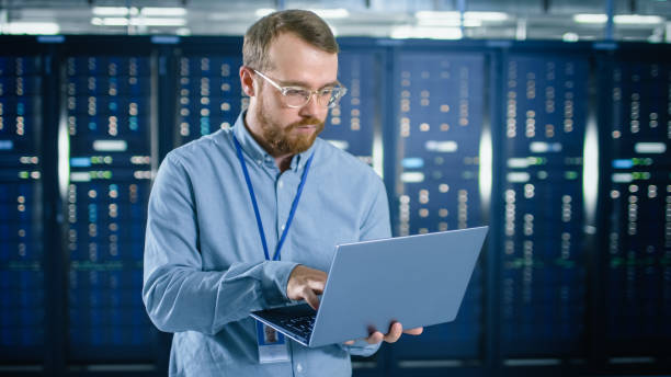 istock Bearded IT Specialist in Glasses is Working on Laptop in Data Center Next to Server Racks. Running Diagnostics or Doing Maintenance Work. 1131198134