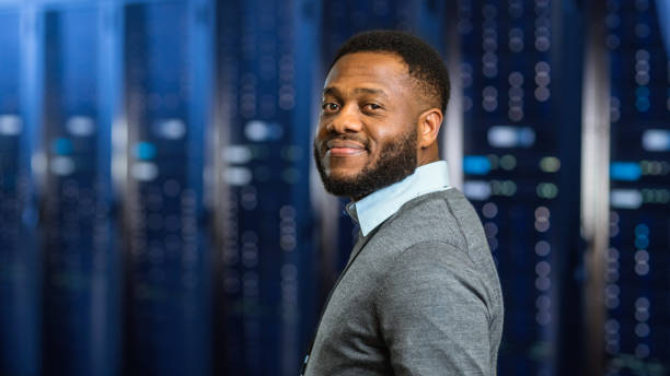 young black data center it technician standing in server rack corridor und smiling to the camera. - network server rack computer black stock-fotos und bilder