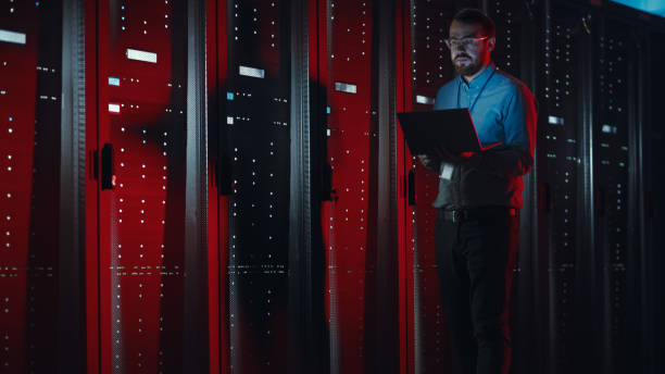 istock Bearded IT Specialist in Glasses is Working on Laptop in Data Center while Standing Near Server Rack. Running Diagnostics, Doing Maintenance Work. Emergency Red Light from Side Illuminating Specialist 1131198141