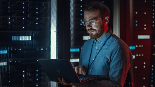 istock Bearded IT Specialist in Glasses is Working on Laptop in Data Center while Standing Near Server Rack. Running Diagnostics, Doing Maintenance Work. Emergency Red Light from Side Illuminating Specialist 1131198157
