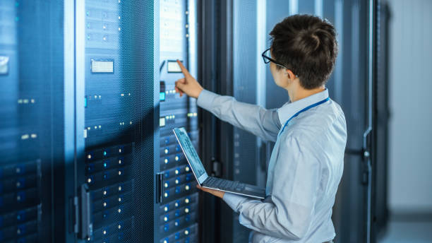 in the modern data center: it engineer standing beside open server rack cabinets, does wireless maintenance and diagnostics procedure with a laptop. he pushes button. - network server rack computer mainframe imagens e fotografias de stock