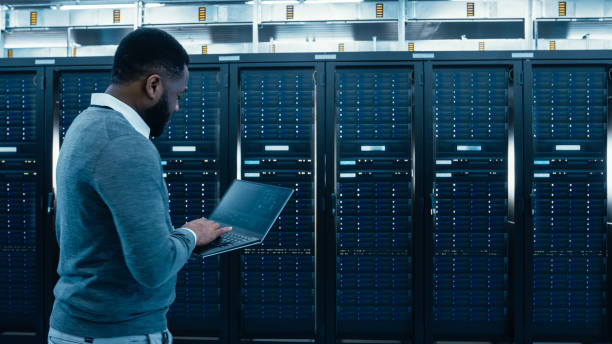 black data center it technician walking through server rack corridor with a laptop computer. he is visually inspecting working server cabinets. - network server rack computer black imagens e fotografias de stock