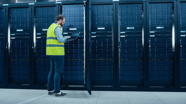 istock IT Engineer in High Visibility Vest is Working on Laptop in Data Center while Standing Before Server Rack. Running Diagnostics or Doing Maintenance Work. 1131198217