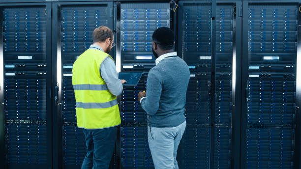 istock IT Administrator with a Laptop Computer in High Visibility Vest and Black Male Engineer are Talking in Data Center while Standing Before a Server Rack. Running Diagnostics or Doing Maintenance Work. 1131198237