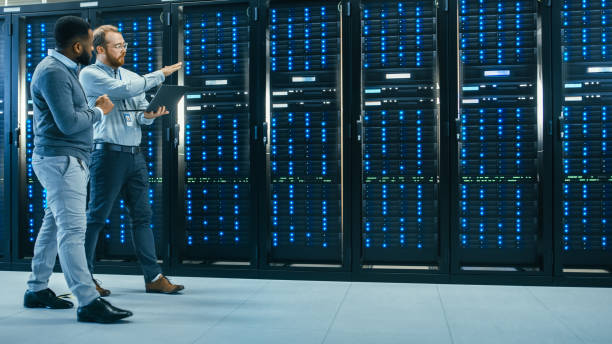 técnico de ti con un ordenador portátil e ingeniero masculino negro colega están hablando en el centro de datos mientras camina al lado de racks de servidores. ejecución de diagnósticos o trabajo de mantenimiento. - data technology network server center fotografías e imágenes de stock