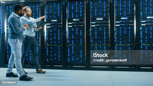 It Technician With A Laptop Computer And Black Male Engineer Colleague Are Talking In Data Center While Walking Next To Server Racks Running Diagnostics Or Doing Maintenance Work Stock Photo - Download Image Now