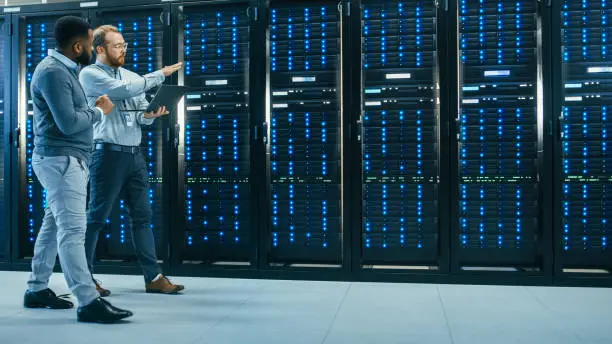 Photo of IT Technician with a Laptop Computer and Black Male Engineer Colleague are Talking in Data Center while Walking Next to Server Racks. Running Diagnostics or Doing Maintenance Work.