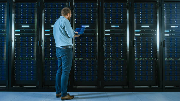 istock Bearded IT Specialist in Glasses is Working on Laptop in Data Center while Standing Before Server Rack. Running Diagnostics or Doing Maintenance Work. 1131198292