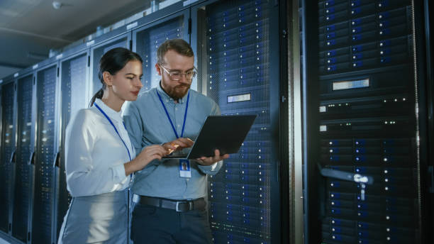 istock Bearded IT Technician in Glasses with a Laptop Computer and Beautiful Young Engineer Colleague are Talking in Data Center while Working Next to Server Racks. Running Diagnostics or Doing Maintenance Work. 1131198315