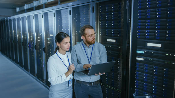 istock Bearded IT Technician in Glasses with a Laptop Computer and Beautiful Young Engineer Colleague are Talking in Data Center while Working Next to Server Racks. Running Diagnostics or Doing Maintenance Work. 1131198316