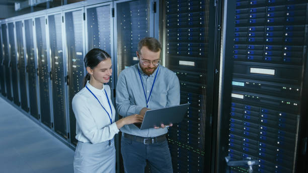 istock Bearded IT Technician in Glasses with a Laptop Computer and Beautiful Young Engineer Colleague are Talking in Data Center while Working Next to Server Racks. Running Diagnostics or Doing Maintenance Work. 1131198318