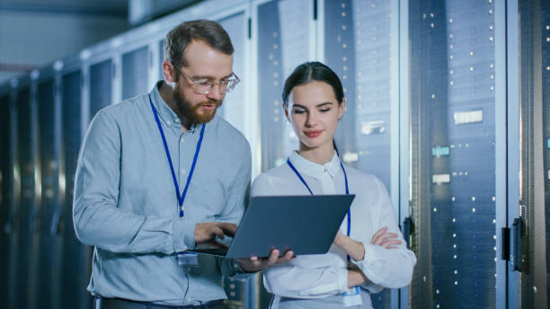 istock Bearded IT Technician in Glasses and Beautiful Young Engineer Colleague with a Laptop Computer are Talking in Data Center while Working Next to Server Racks. Running Diagnostics or Doing Maintenance Work. 1131198325