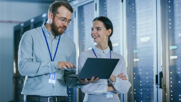 istock Bearded IT Technician in Glasses and Beautiful Young Engineer Colleague with a Laptop Computer are Talking in Data Center while Working Next to Server Racks. Running Diagnostics or Doing Maintenance Work. 1131198334