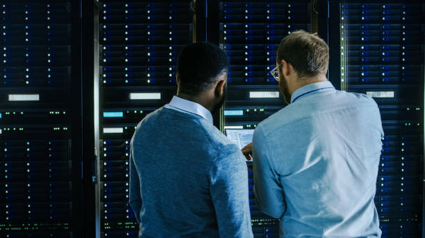 istock Bearded IT Technician in Glasses with a Laptop Computer and Black Male Engineer Colleague are Talking in Data Center while Working Next to Server Racks. Running Diagnostics or Doing Maintenance Work. 1131198355