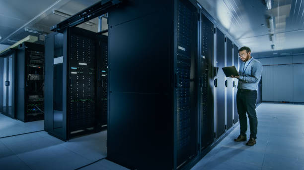 istock Bearded IT Specialist in Glasses is Working on Laptop in Data Center while Standing Before Server Rack. Running Diagnostics or Doing Maintenance Work. 1131198385