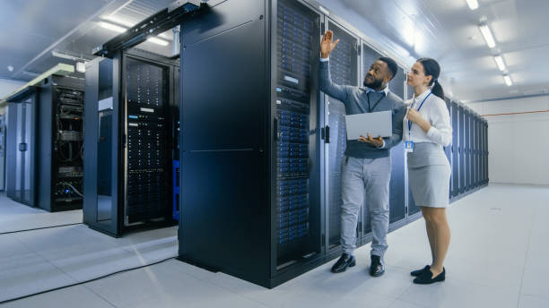 istock Black IT Technician with a Laptop Computer Gives a Tour to a Young Intern. They Talk in Data Center while Walking Next to Server Racks. Running Diagnostics or Doing Maintenance Work. 1131198425
