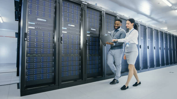 istock Black IT Technician with a Laptop Computer Gives a Tour to a Young Intern. They Talk in Data Center while Walking Next to Server Racks. Running Diagnostics or Doing Maintenance Work. 1131198427