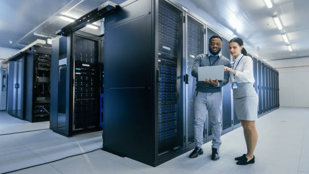 istock Black IT Technician with a Laptop Computer Gives a Tour to a Young Intern. They Talk in Data Center while Walking Next to Server Racks. Running Diagnostics or Doing Maintenance Work. 1131198439