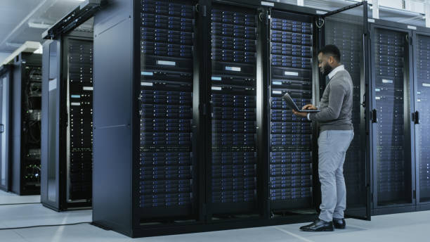 black it specialist is working on laptop in data center while standing before server rack. running diagnostics, putting in data or doing maintenance work. - network server rack computer black imagens e fotografias de stock