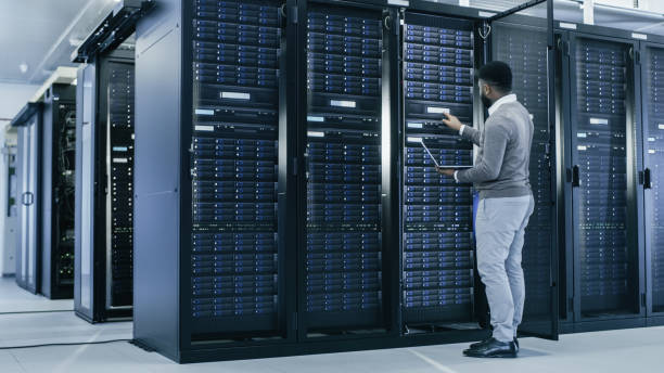 istock Black IT Specialist is Working on Laptop in Data Center while Standing Before Server Rack. Running Diagnostics, Putting in Data or Doing Maintenance Work. 1131198447