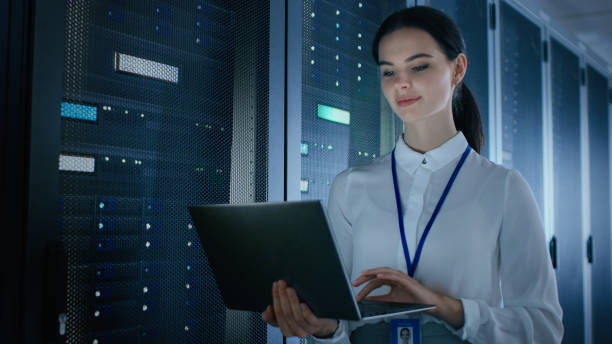 istock Confident and Beautiful Female IT Engineer is Working on Laptop in Data Center while Standing Before Server Rack. Running Diagnostics or Doing Maintenance Work. 1131198452