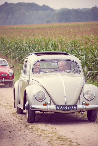 Two vintage Volkswagen cars, a Beetle and Karmann Ghia driving on a country road in between corn fields near the village of Beekbergen in The Netherlands with people inside the cars.