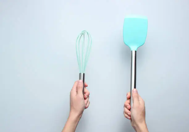 Hand holding silicone shpatula and  whisk for cooking with metal handle on a gray background. Top view.