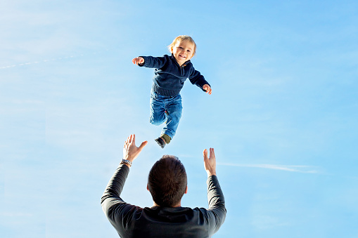 Little toddler boy, flying in the sky, dad throwing him high in the air. Family, enjoying winter view of snowy mountains and frozen lake on a sunny day