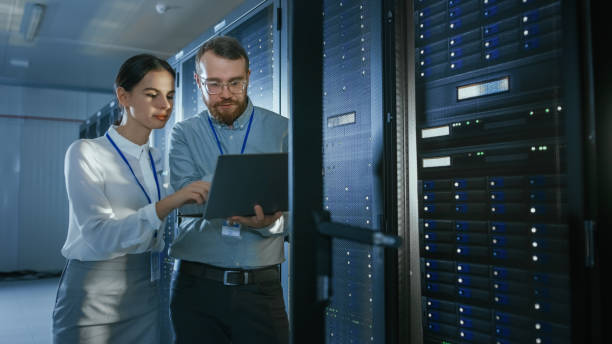 istock Bearded IT Technician in Glasses with a Laptop Computer and Beautiful Young Engineer Colleague are Talking in Data Center while Working Next to Server Racks. Running Diagnostics or Doing Maintenance Work. 1131209052