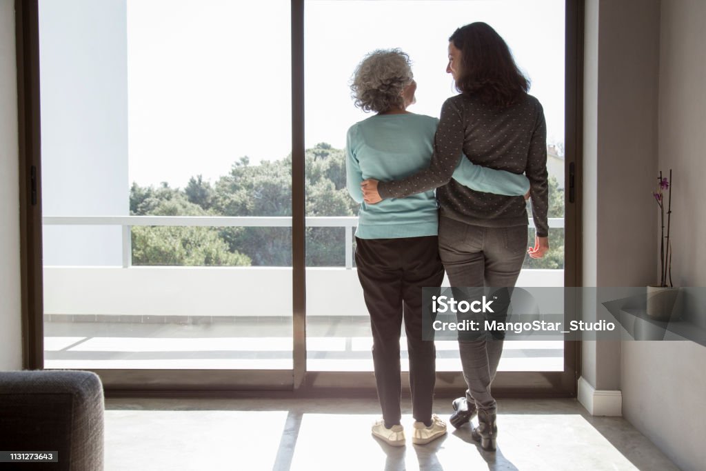 Rear view of happy mother and daughter standing embracing Rear view of happy mother and daughter standing embracing at window. Senior and mid adult women hugging and talking at home. Family relationships concept Senior Adult Stock Photo