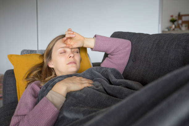 Sick woman lying on grey sofa at home, holding hands on chest