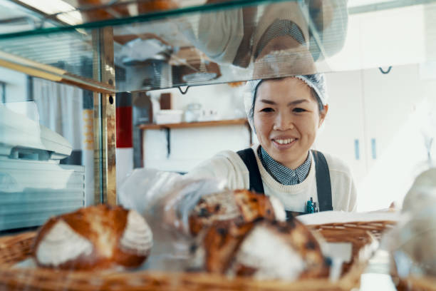 ミッドアダルト女性は、彼女の小さなビジネスベーカリーのディスプレイにパンを入れて - パン屋 ストックフォトと画像