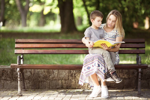 istock Mother and son sitting on a bench outdoors in park and reading a book 1131300110