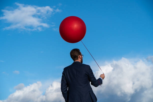 istock Businessman Holding Red Balloon Against Blue Cloudy Sky 1131302450