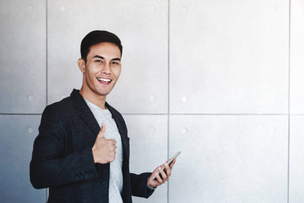 istock Young Happy Businessman Smiling and Show Thumbs Up while Using Smartphone. Standing by the Industrial Concrete Wall. Looking at Camera 1131302521