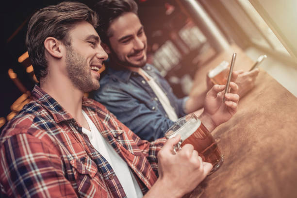 Men in bar Two handsome male friends in bar drinking beer with smartphones in hands and smiling. friends in bar with phones stock pictures, royalty-free photos & images