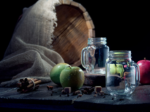 Two mason jars for serving apple cider on the table with fresh apples, spices and a cask on the background