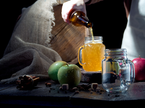 Pouring apple cider served into a mason jar on the table with fresh apples, spices and a cask on the background