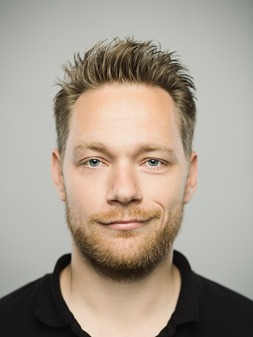 Close up portrait of young adult scandinavian man with happy expression against gray white background. Vertical shot of caucasian real people smirking in studio with blond hair and modern haircut. Photography from a DSLR camera. Sharp focus on eyes.