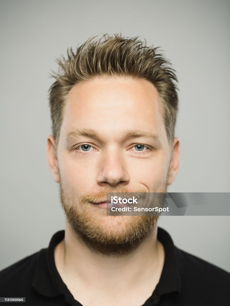 Portrait of real caucasian man with happy expression Close up portrait of young adult scandinavian man with happy expression against gray white background. Vertical shot of caucasian real people smirking in studio with blond hair and modern haircut. Photography from a DSLR camera. Sharp focus on eyes. Men Stock Photo