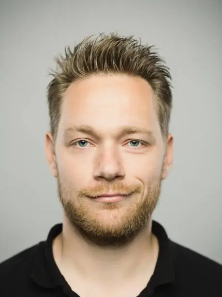 Close up portrait of young adult scandinavian man with happy expression against gray white background. Vertical shot of caucasian real people smirking in studio with blond hair and modern haircut. Photography from a DSLR camera. Sharp focus on eyes.